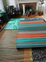 Three outdoor rugs laid on top of each other indoors in front of a fireplace. Bottom rug has green flowers on a yellow border, middle is teal with leaf pattern, top is multicolor striped pattern.