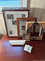 Five large reddish-brown wooden multi-picture frames still wrapped in protective plastic with additional individual frames and shelf beside them on wooden surface near window.