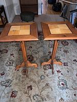 Two wooden decorative tables on a floral rug, showing square tops with mosaic wood tile designs and tripod pedestal bases.