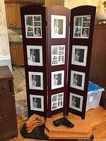 Photo of the 3-panel privacy screen with photo frame inserts, standing upright. Dark wood finish. Multiple black and white photos in frames. One frame has broken glass.