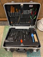 Open black hard case showing multiple hand tools including screwdrivers with amber handles, green pliers, wrenches, hex keys, brush and small tool attachments.