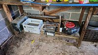 Wide shot of tools displayed on shelves in garage setting, containing large and small toolboxes, hand tools, a black bucket-like container, and other miscellaneous tools scattered around.