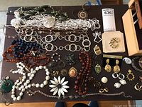 Full view of various costume jewelry pieces spread on a surface including beaded necklaces, pins, earrings, watches, bracelets, and a silver color compact.