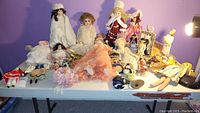 Wide table view showing a variety of vintage dolls in different costumes and poses on a white folding table against a purple wall.