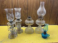 Five lamps displayed on a yellow table with a wood-paneled background: two glass hurricane oil lamps, two electric glass lights with crystals, and a small blue glass oil lamp.