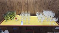 Full lot overview showing green goblets, clear wine glasses (plain and ornate), tall ornate wine glasses, tall goblets, and a Wrightstown Township mug on yellow surface against wood panel background.