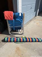 Beach chair folded and standing upright with a red towel draped over, Tommy Bahama umbrella in carrying case on ground