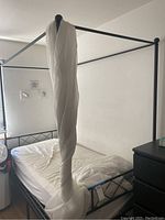 Full view of the assembled black metal canopy bed frame with white fabric loosely draped on one of the posts. Mattress is on the bed frame but optional and not included.