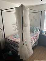 Black metal canopy bed frame with sheer white fabric draped over one vertical post, set up with colorful bedding (not included) against a wall with decorative text.