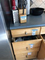 Kitchen counter displaying a wooden knife block holding knives and scissors, metal box grater, and a black bowl. Labels with item number visible.