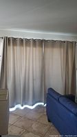Curtain panels covering a large window or sliding glass door. Curtains are beige with grommets along the top edge hung on a curtain rod. Partial view of adjacent furniture and tiled floor.
