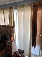 Curtains hanging on a window rod in a living room setting, showing light cream vertical striped fabric with grommet tops.