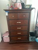 Front view of dark wooden highboy dresser, showing six drawers with metal handles and various personal items on top.