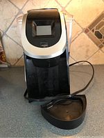 Front view of Keurig K-Select coffee maker showing black and silver design, digital display, drip tray, and power cord.