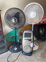 Photo showing the Lasko Motion Heat Plus digital ceramic heater below two oscillating floor fans, one black and one white, against a background with a tire and other items.