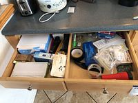 Two open drawers on a kitchen counter containing various office supplies such as reams of copy paper, boxes of binder clips, tape rolls, binders, paper clips, small tools, and pouches.