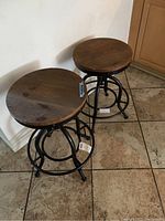 Two wooden and metal bar stools side by side, showing round wooden seats and black metal bases with circular footrests on tiled floor.