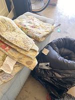 Quilt folded on a light-colored cushion next to a rolled sleeping bag in a black plastic bag, with part of a bicycle visible in the background.