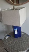 Blue ceramic table lamp on round table with beige rectangular fabric shade, metal finial at top, tag on lamp base