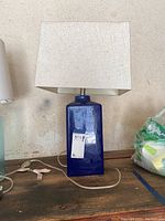 Full view of blue ceramic table lamp with white rectangular fabric lampshade on wooden surface against neutral background.