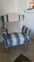 Full view of the striped upholstered armchair with wingback design, wooden legs, and two pillows displayed on seat.