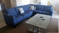 Full view of L-shaped blue upholstered sectional sofa with tufted cushioning and two attached pieces. Sofa sits in a living room on a brown carpet.