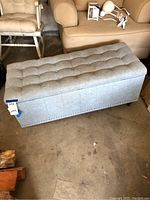 Side view of rectangular light gray button tufted upholstered storage ottoman, showing nailhead trim and missing legs condition.