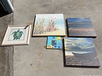 Five pieces of wall art laid out on garage floor including turtle watercolor framed painting, coastal prints, and small colorful mirror.