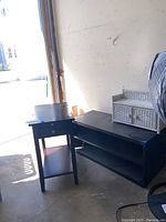 Photo showing three furniture items grouped: white wicker shelf with two drawers on top of TV stand, black wooden end table to left side, all placed in garage space with concrete floor and white wall.
