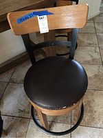 Close-up photo of one swivel bar stool showing wooden backrest, worn black vinyl seat peeling at edges, metal frame and footrest, tile floor background.