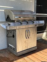 Left-front angle view of the stainless steel barbecue showing the grill body, lid, control knobs, and storage cabinet below.