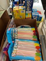 Photo showing various household disposable items and storage products arranged in an open cardboard box on a counter.