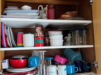 Photo showing two cabinet shelves with numerous kitchenware items including platters, mugs, glasses, plastic kids cups, candle holders, and a glass coffee carafe.