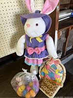 Large plush bunny in purple outfit holding Happy Easter wooden sign with a bag of colorful plastic eggs on floor next to it.