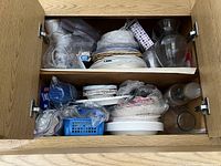 Two-shelf wooden cabinet with assorted disposable party supplies including paper plates, plastic utensils, napkins, cups, and some glassware on upper and lower shelves.