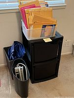 Overview photo showing white mesh basket filled with assorted mailing envelopes on top of a black two-drawer plastic filing cabinet, blue tarp folded beside it, black trash bin with white power strip inside beside filing cabinet.