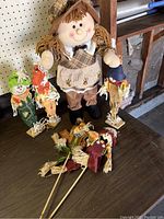 Five fall-themed scarecrow decorations arranged on a surface including a large fabric standing scarecrow doll and four small wooden scarecrow stakes with colorful hats and straw details.