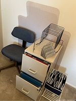 Photo showing beige two drawer file cabinet with top drawer partially open revealing manila hanging folders. Bottom drawer open revealing blue hanging folders. Black metal wire file organizers on top of the cabinet. Black office chair with fabric upholstery beside cabinet. Transparent plastic desk mat behind cabinet and chair.