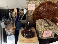 Photo shows a collection of vintage kitchen items on a stove and countertop, including Lucite tray with original box, amber Pyrex bowls on a wooden cutting board, a wooden block with various knives and tools, and a blue crock with utensils.