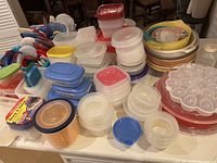 Large collection of assorted plastic food storage containers with lids arranged on a surface, including rectangular and round shapes, and some with colored lids mostly blue, red, and white.
