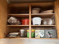Cupboard shelf showing small appliances and baskets including rice cooker, hand mixer in box, glass baking dish, multiple wicker baskets