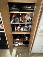 Full view of cabinet door open showing all four shelves packed with kitchen items including baking sheets, glassware, pots, pans, and kitchen utensils.