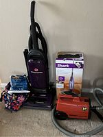 Photo showing the upright Hoover WindTunnel Lite vacuum, boxed Shark steam mop, orange Eureka Mighty Mite canister vacuum, and tote bag with vacuum bags and accessories.