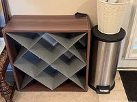 Photo showing a wooden wine cabinet with silver metal diamond-shaped compartments and two garbage cans—one stainless steel step-on type on the floor and one small white woven basket type on a black round table.