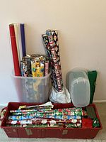 Multiple rolls of assorted gift wrap standing inside a white plastic container, with more rolls placed in a red plastic bin on the floor, plus two plastic storage boxes on the right.