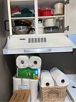 Upper kitchen cabinet open, showing Pyrex glass measuring cup, plastic containers including a red microwave-safe pot, and white metal mixing bowls. Below the cabinet, rack holds multiple rolls of paper towels and a box of flushable wipes.