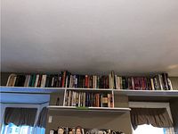 Books shelved on a high shelf, a mix of hardcover and paperback in a room with window curtains.