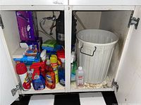 Photo showing inside cabinet with assorted cleaning sprays, a large gray trash bin, sponges, and other cleaning supplies.