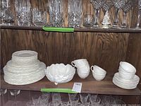 Full view of stacked plates, cups, and footed bowl on a wooden shelf with crystal glassware above.