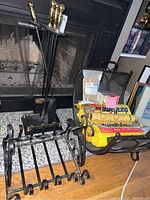Two metal log holders placed on floor in front of fireplace, one flat with multiple slots and one with curved bars. Nearby are Duraflame fire logs and fire-starting items.
